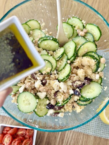A bowl of Greek couscous salad featuring sliced cucumbers, chickpeas, feta cheese, and olives, with a drizzle of dressing being added from a small dish.