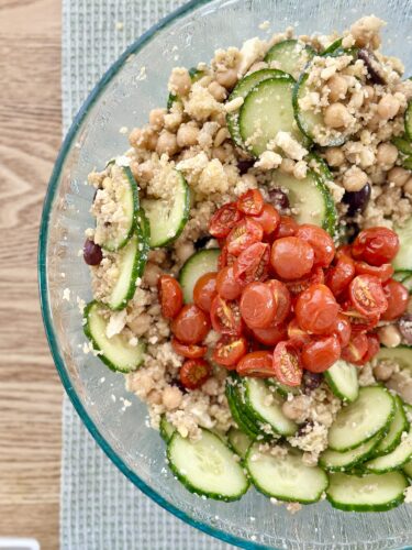 A bowl of Greek couscous salad featuring sliced cucumbers, chickpeas, Kalamata olives, and cherry tomatoes, with a light, textured appearance.