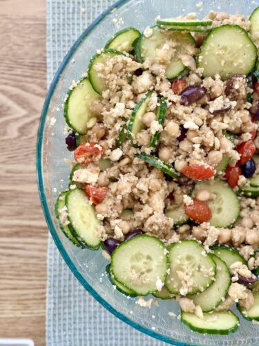 A bowl of Greek couscous salad featuring sliced cucumbers, cherry tomatoes, chickpeas, Kalamata olives, feta cheese, and herbs, arranged on a light blue placemat.