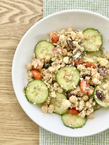 A bowl of Greek couscous salad featuring chopped cucumbers, cherry tomatoes, chickpeas, Kalamata olives, and crumbled feta cheese, with a light dressing.