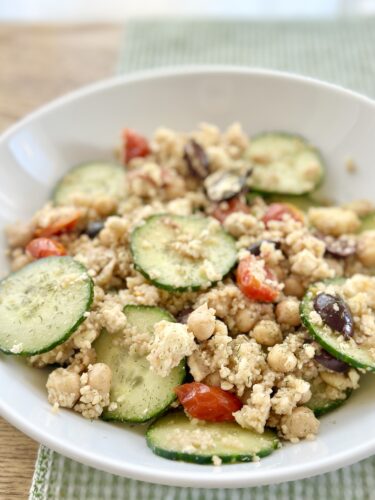 A serving of Greek couscous salad featuring sliced cucumbers, cherry tomatoes, chickpeas, Kalamata olives, and crumbled feta cheese in a white bowl.