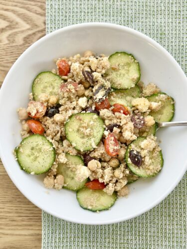 A bowl of Greek couscous salad featuring sliced cucumbers, cherry tomatoes, chickpeas, Kalamata olives, and crumbled feta cheese, garnished with herbs.