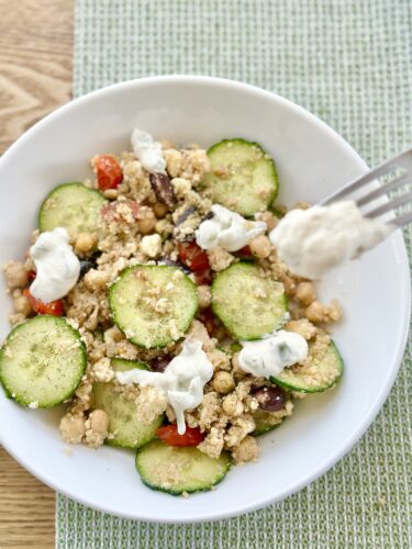 A bowl of Greek couscous salad featuring sliced cucumbers, cherry tomatoes, chickpeas, Kalamata olives, crumbled feta cheese, and a drizzle of tzatziki sauce, presented on a light green placemat.