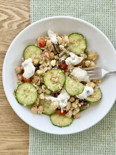 A bowl of Greek couscous salad featuring sliced cucumbers, chickpeas, Kalamata olives, cherry tomatoes, and crumbled feta cheese, drizzled with tzatziki sauce and served on a light green placemat.