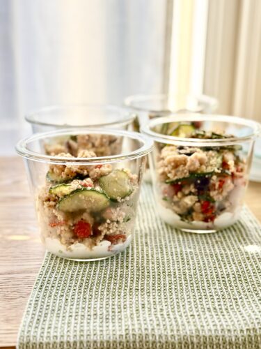 Glass jars filled with Greek couscous salad featuring cucumbers, peppers, and dressing, arranged on a textured green napkin.