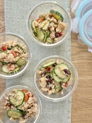 Containers filled with Greek couscous salad featuring cucumbers, cherry tomatoes, chickpeas, and Kalamata olives, placed on a woven mat next to food storage lids.