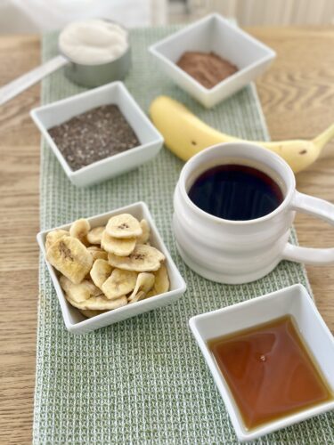 A wooden table with a green placemat featuring small bowls containing chia seeds, cocoa powder, banana chips, and a cup of coffee, with a banana and a spoonful of yogurt in the background.