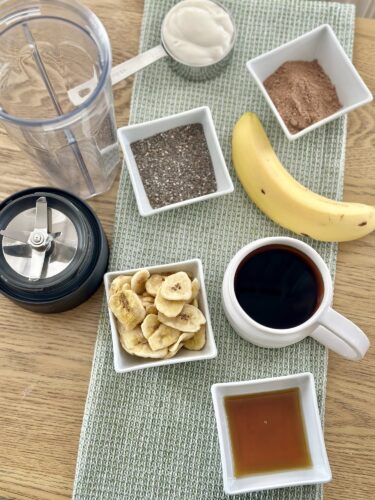A collection of smoothie ingredients arranged on a green cloth, including a blender, yogurt, cacao powder, chia seeds, a banana, dried banana slices, a cup of coffee, and honey.