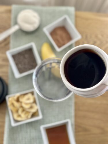 A top-down view of a table setting featuring a white cup filled with dark liquid, surrounded by various containers of ingredients including sugar, cocoa, chia seeds, and dried banana slices on a green fabric mat.