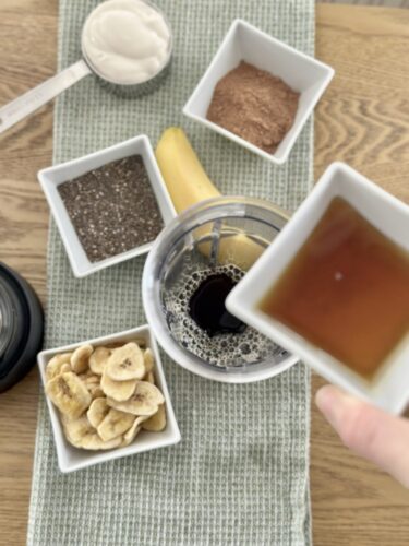A flat lay of ingredients for a smoothie, including banana, chia seeds, yogurt, cocoa powder, dried banana slices, and maple syrup, arranged on a green cloth.