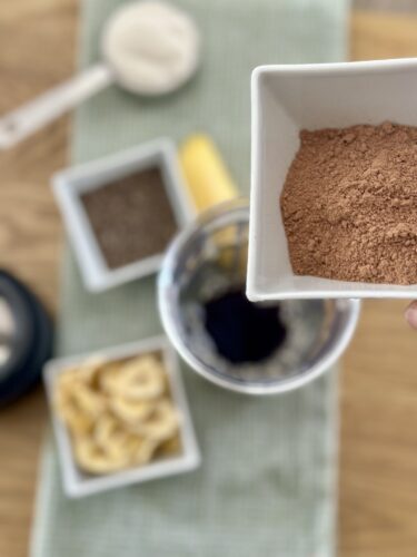 A person holding a small square dish filled with cocoa powder, with various ingredients like dried banana slices, coffee grounds, and a measuring spoon in the background.