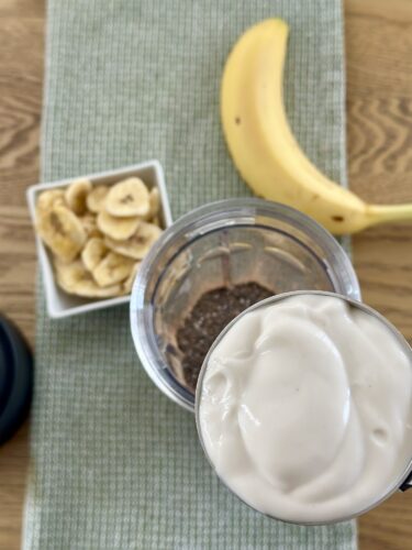 A banana, a bowl of dried banana slices, and a blender containing a creamy smoothie on a green placemat.