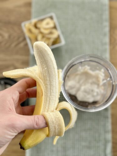 A hand holding a peeled banana in front of a smoothie maker filled with a creamy mixture, with a bowl of banana slices in the background.
