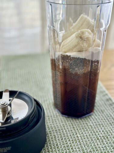 A blender filled with layers of ingredients including bananas, chia seeds, and a chocolate mixture on a green textured mat, with a blender base in the foreground.