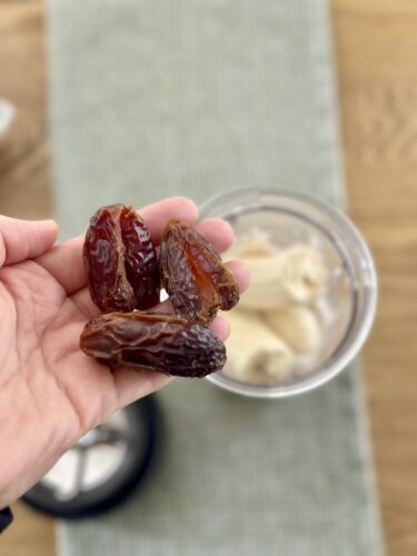 A hand holding three dates with a blender containing banana in the background.