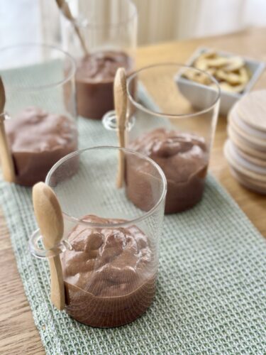 Four cups of chocolate mousse with wooden spoons on a table, with a green placemat and a bowl of snacks in the background.