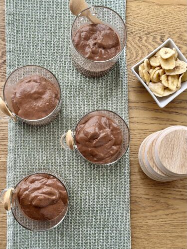 Four glasses of chocolate mousse on a textured green table runner, accompanied by a small bowl of banana chips and a wooden spice container.