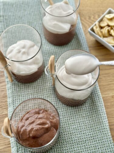 A table setting with four glass cups of dessert, two filled with chocolate mousse topped with whipped cream, one containing a layer of chocolate mousse, and a small bowl of banana chips in the background.