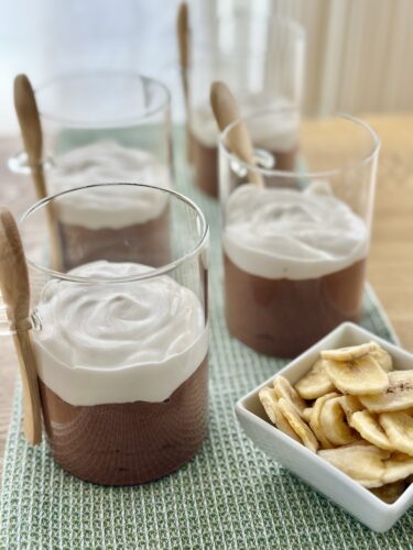 Four glasses of chocolate mousse topped with whipped cream, placed on a textured green mat, accompanied by a small bowl of banana chips.
