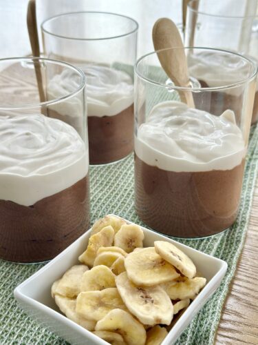 Four dessert glasses filled with chocolate mousse topped with whipped cream, accompanied by a small bowl of banana chips.