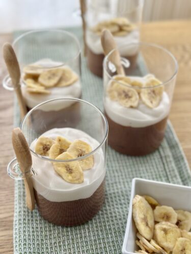 Dessert cups with chocolate mousse topped with whipped cream and banana chips, served with small wooden spoons on a textured green placemat.