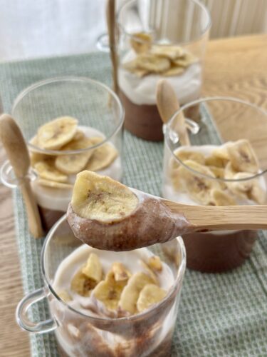 A close-up of a glass dessert with layers of chocolate mousse and yogurt, topped with banana slices. In the background, several similar glasses are arranged on a wooden table.