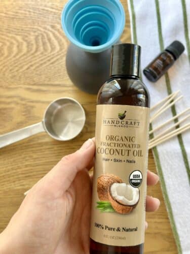 A hand holding a bottle of organic fractionated coconut oil on a wooden surface, with a measuring cup, a funnel, and essential oil in the background.