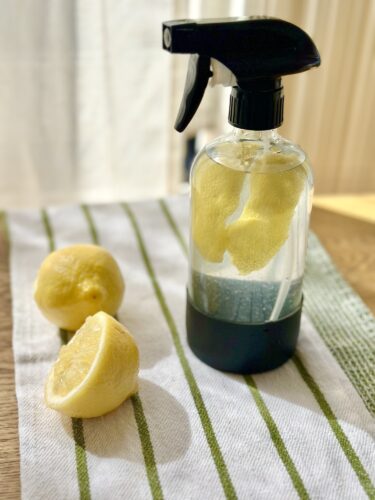A clear spray bottle filled with water and lemon slices, placed on a striped towel with two whole lemons beside it.