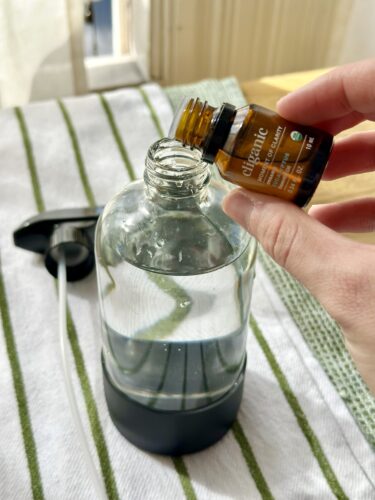 A person pours essential oil from a brown bottle into a clear spray bottle filled with liquid, placed on a striped cloth.