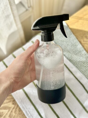 A person holding a clear spray bottle filled with water and bubbles, with a black nozzle, set against a striped cloth background.