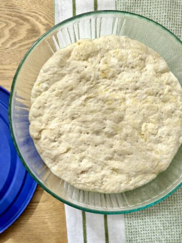 A glass bowl containing risen dough, showing a smooth, slightly puffy surface, on a wooden table.