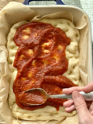 A person spreading tomato sauce on a pizza dough in a baking dish lined with parchment paper.