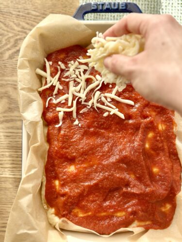 A hand sprinkling shredded cheese over a tray of pizza sauce spread on dough lined with parchment paper.