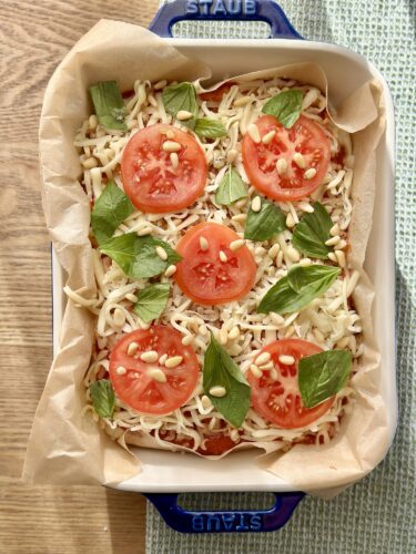 Baking dish with layers of pasta topped with tomato slices, fresh basil leaves, and pine nuts, wrapped in parchment paper.