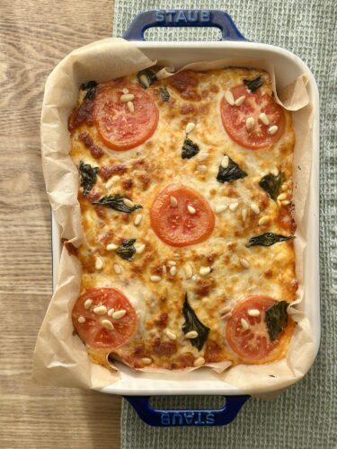 Baked dish with melted cheese, sliced tomatoes, pine nuts, and fresh basil, served in a white casserole dish.