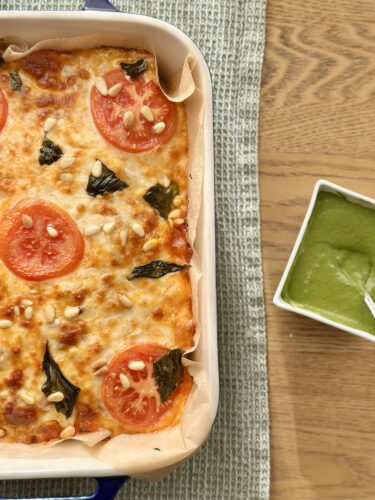 Baked pizza topped with tomatoes, basil, pine nuts, and melted cheese, served alongside a small bowl of green sauce.