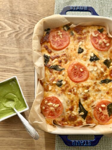 A freshly baked pizza topped with sliced tomatoes, pine nuts, and basil, served in a blue Staub baking dish with a side of green sauce.