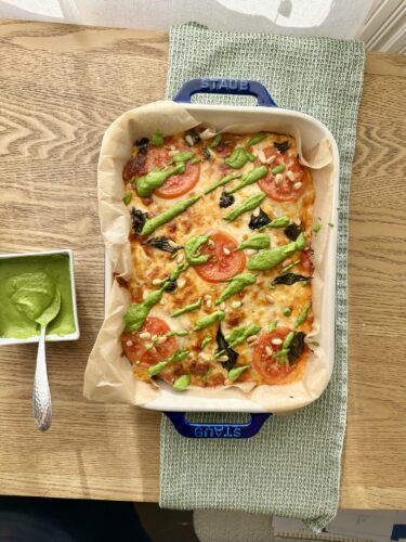 Dish of baked pizza topped with tomatoes, green herbs, and drizzled with sauce, placed on a wooden table next to a small bowl of green sauce.