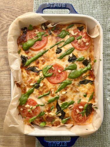 A baked dish featuring a golden crust topped with melted cheese, fresh tomato slices, green basil, and drizzles of vibrant pesto sauce, served in a white baking dish lined with parchment paper.