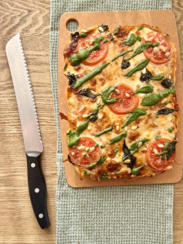 A freshly baked pizza topped with slices of tomato, spinach, and a drizzle of pesto, placed on a wooden cutting board with a serrated knife beside it.