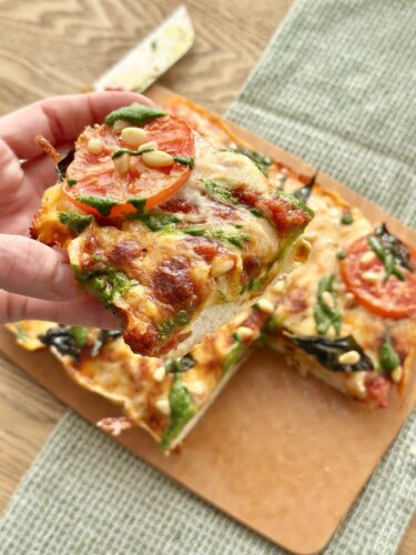 A hand holding a slice of vegetable and cheese pizza topped with tomatoes and pine nuts, with more pizza on a wooden cutting board in the background.