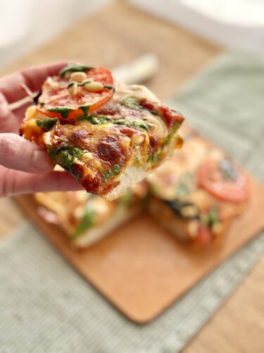 A hand holding a slice of vegetable pizza topped with tomatoes and cheese, with additional slices in the background on a wooden board.