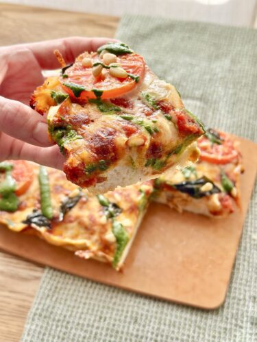 A hand holding a slice of vegetable pizza topped with tomatoes, spinach, and pine nuts, with the pizza resting on a wooden board.