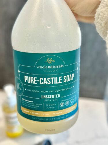 A bottle of Whole Naturals Pure-Castile soap labeled as unscented, showcasing a clear liquid with a vibrant turquoise label. The bottle is held by a hand and is displayed against a blurred background.