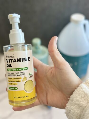A hand holding a bottle of Yoken Vitamin E Oil, labeled as 100% pure and natural, with a focus on skin, face, and nails.