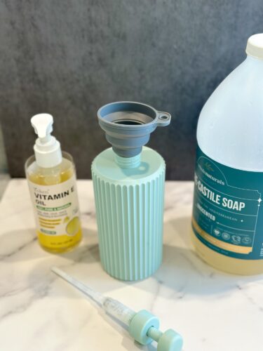 A collection of cleaning and skincare products including a bottle of castile soap, a bottle of vitamin E oil, a silicone funnel, and a dropper, all on a marble surface.