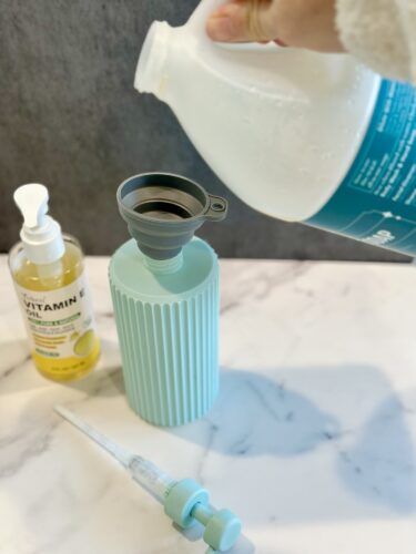 A person pouring a liquid from a transparent bottle into a teal ribbed container using a funnel, with a bottle of vitamin oil and a dropper nearby.