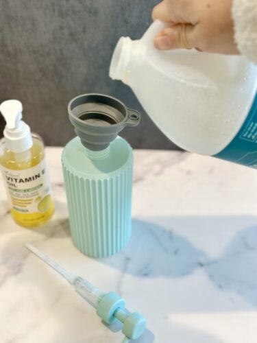 A person pouring liquid from a bottle into a light blue container equipped with a funnel, alongside a dropper and a bottle of vitamin oil.