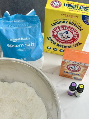 A bowl filled with white powdery substance surrounded by various cleaning products including a blue bag of Epsom salt, a yellow box of Arm & Hammer laundry booster, a red box of baking soda, and small bottles of essential oils.