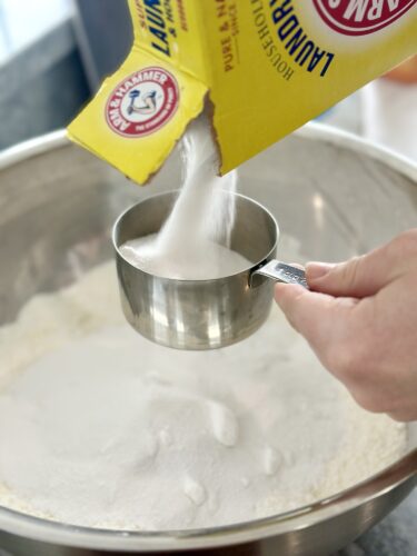 A hand pouring baking soda from a yellow box into a measuring cup above a silver mixing bowl filled with flour.